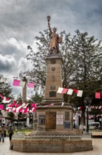Statue on a stone pedestal in a plaza with red and white flags and trees, under a cloudy sky,