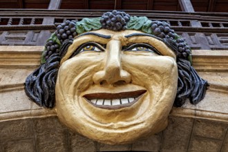 Stone mask with smiling face on an architectural structure, surrounded by green and brown tones,