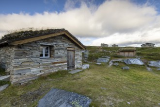 A small stone hut with a grass roof on a rocky meadow, surrounded by other wooden huts and sparse