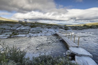 A small wooden bridge crosses a wide river, surrounded by rocks and sparse vegetation in the