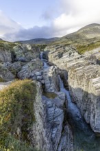 Deep gorge with meandering watercourse, surrounded by jagged rocks and mountains under a cloudy