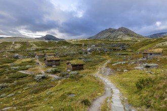 Wooden huts in an open meadow in a remote mountain landscape, with winding paths and clouds in the