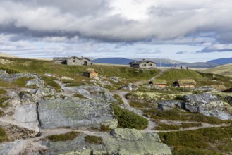 Various wooden cabins on a rocky plain, surrounded by grass and mountains under a cloudy