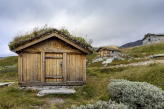 Wooden huts with grass-covered roofs in a hilly, rural landscape under a cloudy sky, Peer Gynt