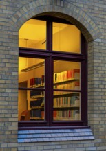 A view through the window of a library, shelves full of books illuminated by a warm, yellow light,