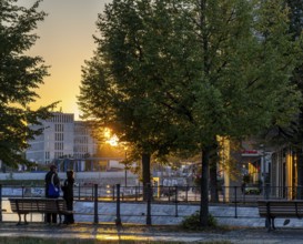 A sunset scene in Berlin with two people standing near a river, surrounded by trees and urban