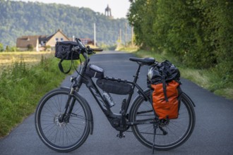 An electric bicycle with an orange bag stands on a rural road, surrounded by green nature and a