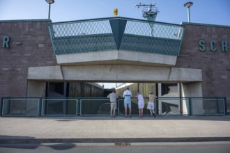Four people stand in front of a modern lock structure made of concrete and blue railings, Minden