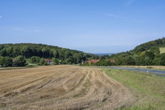 View of a harvested field with neighbouring village and wooded hills under a clear sky, Halle,