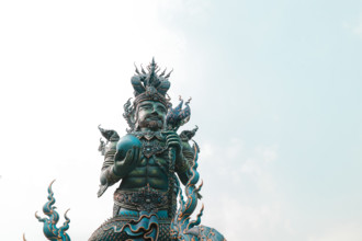 A statue of a mythical figure with intricate details against a pale sky in Wat Rong Suea Ten or