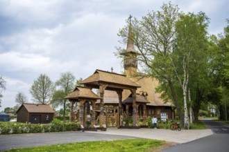 Wooden church with characteristic carvings, along a country road, surrounded by trees and sheds,