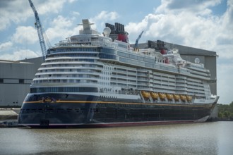 A large cruise ship lies calmly in the water next to a large shipyard on a partly cloudy day,