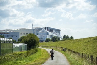A cyclist on a country road next to a shipyard with a cruise ship in the background, cruise ship at