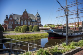 An old ship in front of an impressive brick building, surrounded by manicured nature and water, The