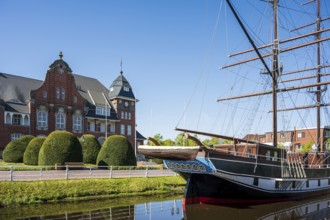 A sailing ship lies in front of a historic building with a blue sky and well-tended bushes on the