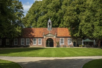 A brick building with a bell tower in front of a grassy area and surrounded by tall trees in the