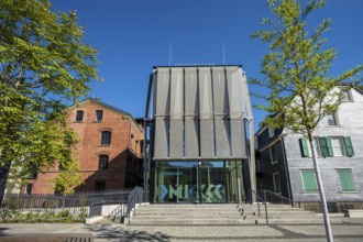 Modern glass building between old houses, surrounded by trees, bright blue sky, Museum of Early