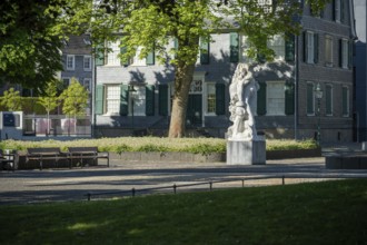 Small square with stone statue in front of an old house with green shutters, illuminated by