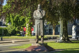 Stone statue of a man with crossed arms in an urban park with surrounding buildings, statue of