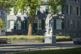 Stone figure in front of a historic building with green shutters, surrounded by blossoming trees,