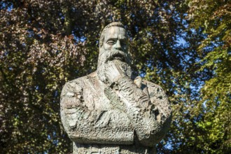 Close-up of a stone statue of a man with a beard, surrounded by trees and a clear sky, statue of