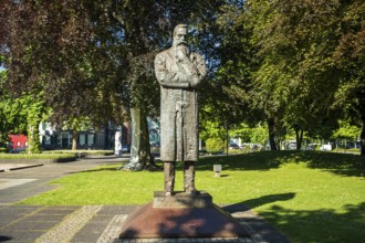 Stone statue of a man with crossed arms in a sunny park with many trees, statue of Friedrich Engels