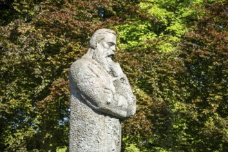 Stone statue of a pensive man surrounded by lush green trees in a park, statue of Friedrich Engels