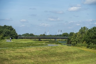 A bridge leads over green areas with trees and a path under a blue sky with few clouds. Stadium