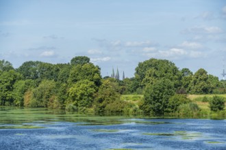 A river with green foliage, church towers in the background and blue sky, Weser cycle path, Weser,