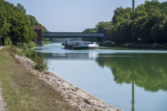 Cargo ship under a bridge, flanked by trees. Tranquil river landscape with greenery, Dortmund-Ems