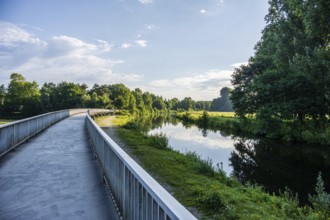 Path along a calm river, lush vegetation and cloudy sky are reflected in the water, Emssee near