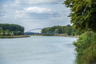 View of a canal with a modern blue bridge under a cloudy sky, surrounded by green trees, Senden,
