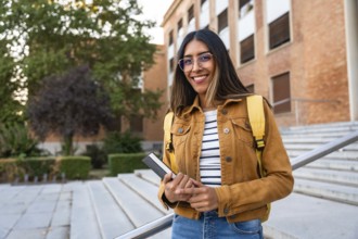 Young woman smiling, wearing glasses and a backpack, holding a book, and standing on university