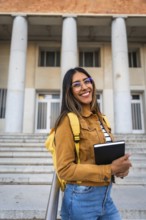 Young woman smiling, wearing glasses and a backpack, standing with a notebook in front of a