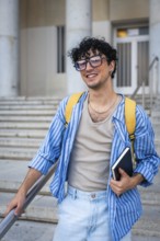 Young man with a backpack and a notebook, smiling while walking up the modern building stairs,