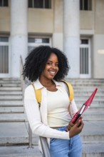 African american woman smiling while holding a notebook, wearing a backpack and standing in front