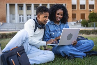 Young black students sitting on green grass, working on a laptop, discussing school project or