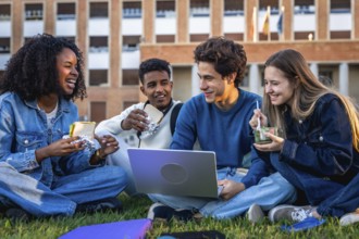 Group of happy young university students from different ethnicities eating, laughing, and using a