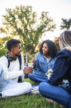 Diverse group of university students sitting on campus grass, chatting, sharing snacks and laughing