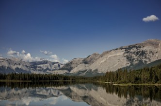 Reflections of rugged mountains of the Canadian Rocky Mountains in a calm lake framed by dense
