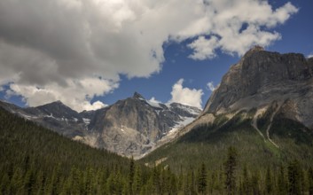 Rocky peaks of the Canadian Rocky Mountains under scattered clouds with a lush forest at the base