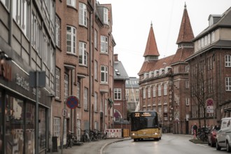 A yellow bus navigates a narrow city street lined with red brick buildings on a gray day in Aarhus,