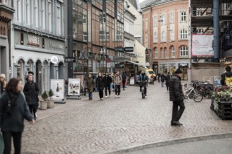 People walk along a bustling cobblestone street lined with various shops in Aarhus, Denmark,