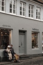A couple sits on a bench outside a gray storefront having a conversation in Aarhus, Denmark,