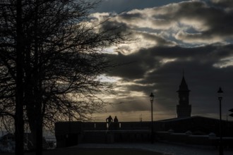 Silhouette scene with tree, lampposts, and tower against a dramatic cloudy sky at sunset in