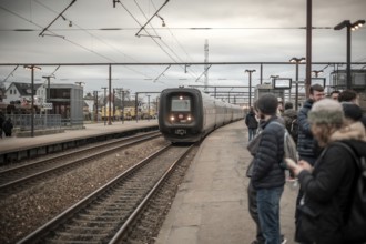 Commuters waiting at a platform as a train approaches, set against an overcast sky in Copenhagen,