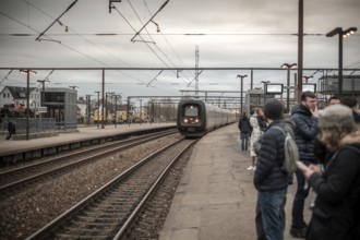 Commuters waiting on a platform as a train approaches at an overcast train station in Copenhagen,