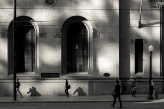 Black and white urban scene with people walking past a sunlit building casting strong shadows in
