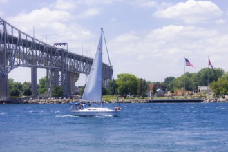 Sailboat gliding near the Blue Water Bridge, connecting Canada and the United States, with flags