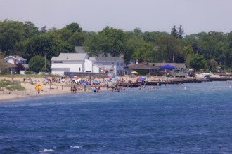A busy beach scene with people enjoying the sun, umbrellas, and nearby houses along the St. Clair